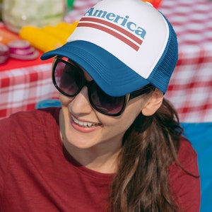 Op de afbeelding: Een vrouw draagt een blauwe en witte trucker-pet met het woord "America" in rode letters. Ze glimlacht en draagt een zonnebril.