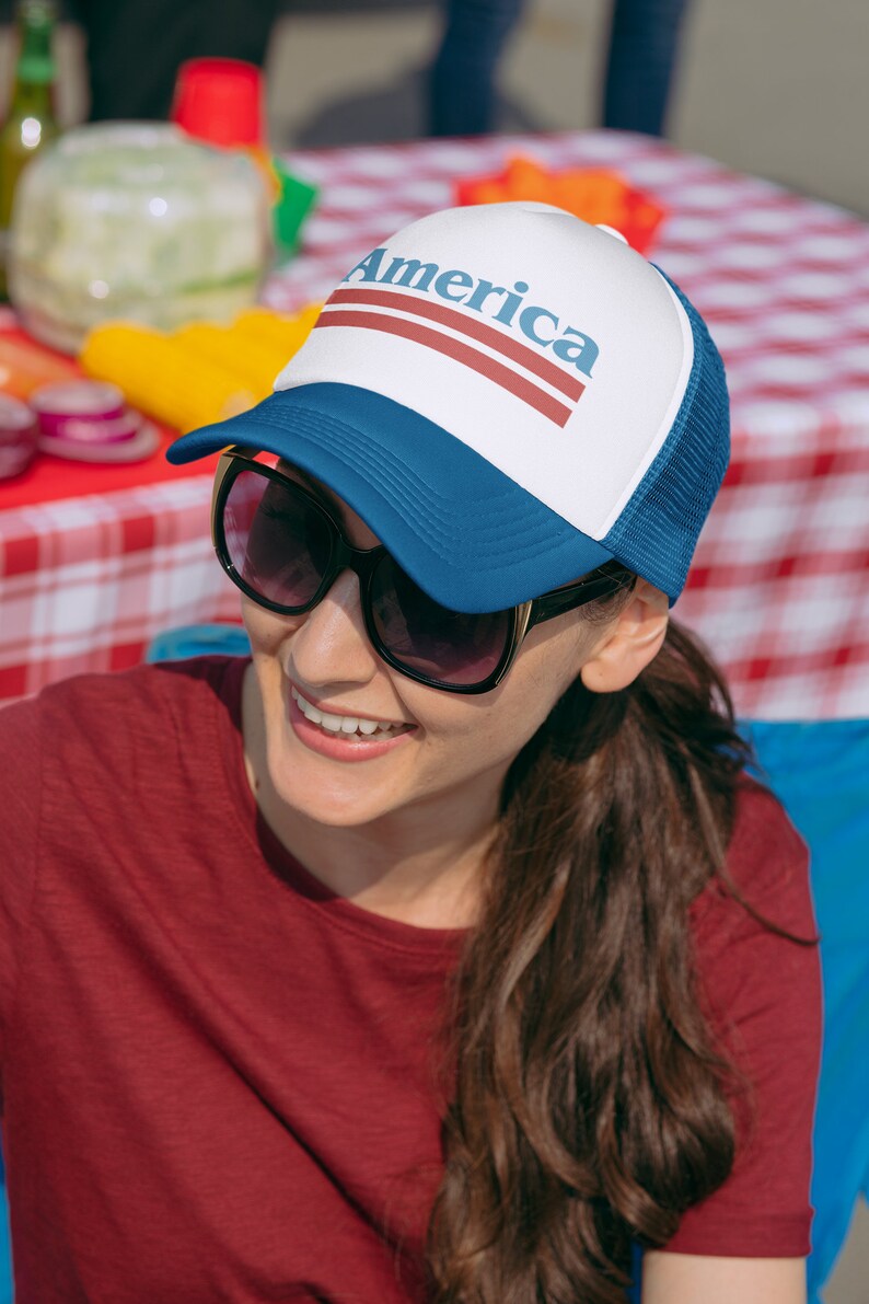 Op de afbeelding: Een vrouw draagt een blauwe en witte trucker-pet met het woord "America" in rode letters. Ze glimlacht en draagt een zonnebril.