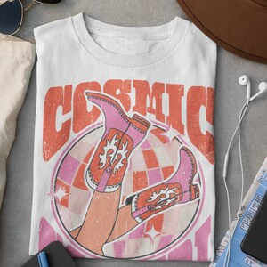 May include: White t-shirt with a retro graphic design featuring a pair of pink cowboy boots with orange accents, a disco ball, and the text "COSMIC"