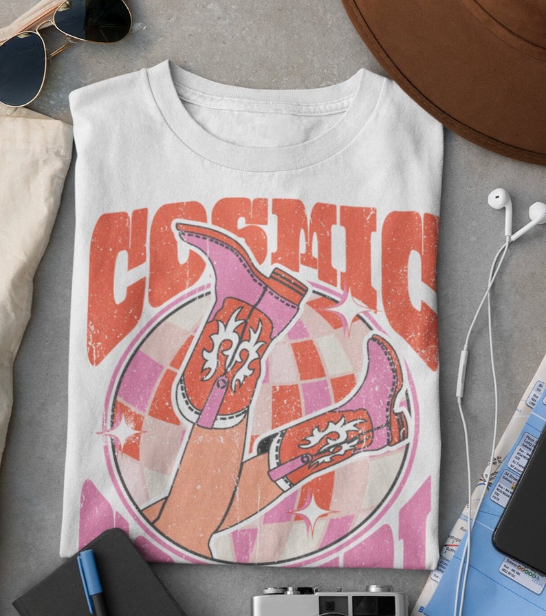 May include: White t-shirt with a retro graphic design featuring a pair of pink cowboy boots with orange accents, a disco ball, and the text "COSMIC"