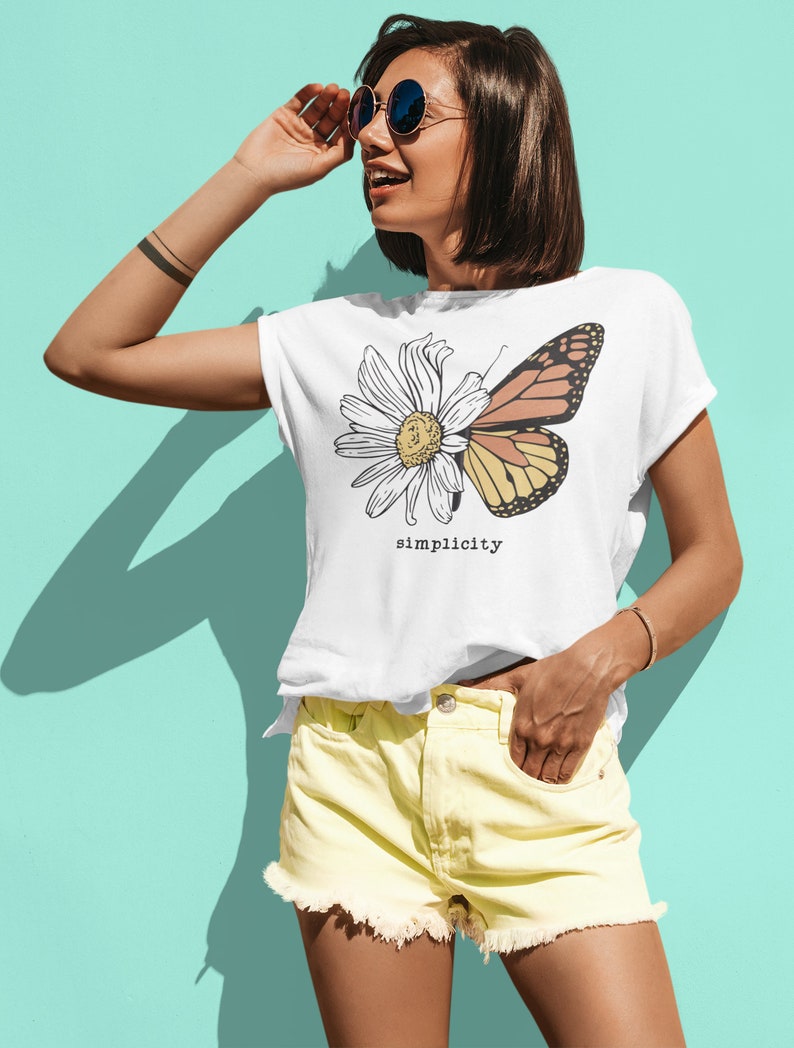 K&ouml;nnte beinhalten: Wei&szlig;es T-Shirt mit einem Motiv einer G&auml;nsebl&uuml;mchen und eines Schmetterlings mit dem Wort "simplicity" darunter gedruckt. Das Model tr&auml;gt eine gelbe Jeans-Shorts und eine Sonnenbrille.