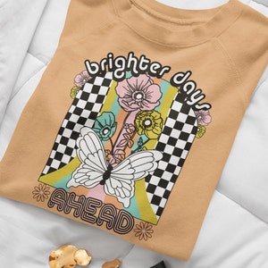 Puede incluir: Una sudadera de color marr&oacute;n claro con un dise&ntilde;o gr&aacute;fico que incluye una mariposa, flores y un patr&oacute;n de cuadros. El texto "brighter days ahead" est&aacute; impreso en la sudadera.