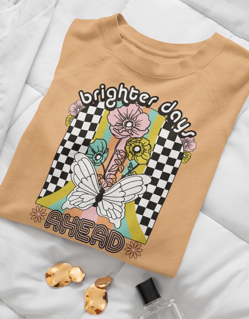 Puede incluir: Una sudadera de color marr&oacute;n claro con un dise&ntilde;o gr&aacute;fico que incluye una mariposa, flores y un patr&oacute;n de cuadros. El texto "brighter days ahead" est&aacute; impreso en la sudadera.