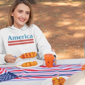 Op de afbeelding: Een witte hoodie met het woord "America" bedrukt in blauwe en rode strepen. De hoodie wordt gedragen door een persoon die aan een tafel zit met een rood-wit-blauw gestreept tafelkleed. Er zijn corn dogs op een bord en een rode beker met sterren op de tafel.