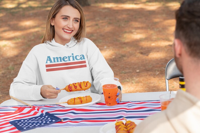 Op de afbeelding: Een witte hoodie met het woord "America" bedrukt in blauwe en rode strepen. De hoodie wordt gedragen door een persoon die aan een tafel zit met een rood-wit-blauw gestreept tafelkleed. Er zijn corn dogs op een bord en een rode beker met sterren op de tafel.