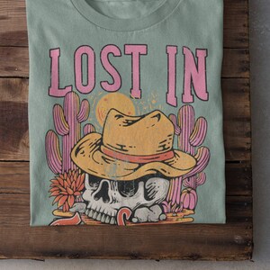 Peut inclure: Un t-shirt vert clair avec le texte "LOST IN" en lettres roses. Le motif repr&eacute;sente un cr&acirc;ne de dessin anim&eacute; portant un chapeau de cowboy et entour&eacute; de cactus.