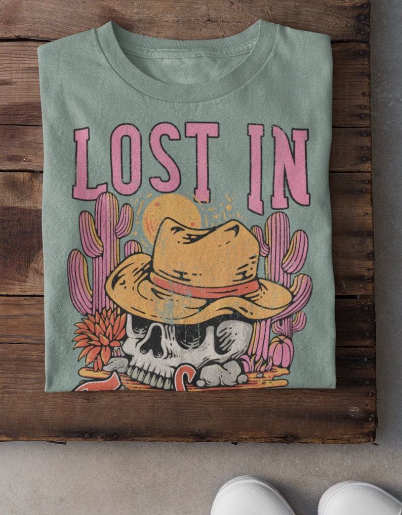 Peut inclure: Un t-shirt vert clair avec le texte "LOST IN" en lettres roses. Le motif repr&eacute;sente un cr&acirc;ne de dessin anim&eacute; portant un chapeau de cowboy et entour&eacute; de cactus.