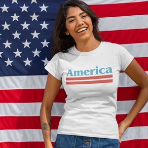 Op de afbeelding: Een wit T-shirt met het woord "America" bedrukt in blauwe en rode letters met twee rode strepen eronder. De persoon die het shirt draagt staat voor een Amerikaanse vlag.