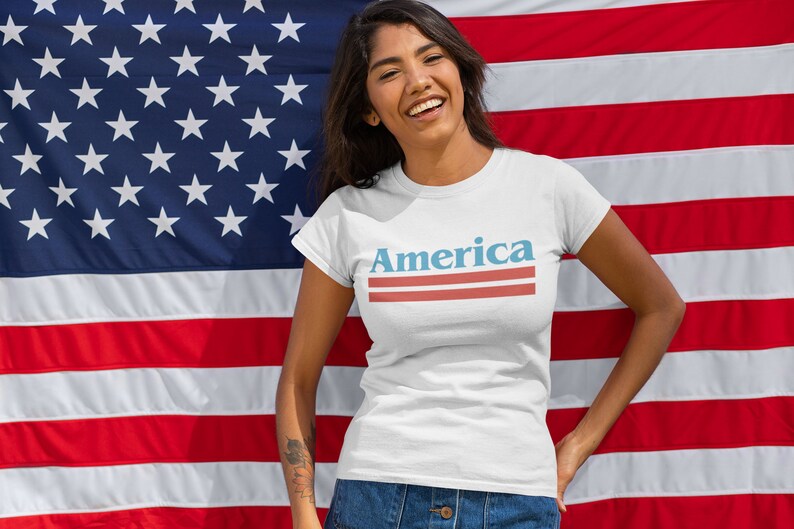 Op de afbeelding: Een wit T-shirt met het woord "America" bedrukt in blauwe en rode letters met twee rode strepen eronder. De persoon die het shirt draagt staat voor een Amerikaanse vlag.