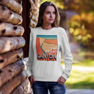 May include: A white sweatshirt featuring a graphic of the Grand Canyon in orange, blue, and beige. The text "THE GRAND CANYON" is printed in a vintage style. The sweatshirt is long-sleeved and worn by a person.