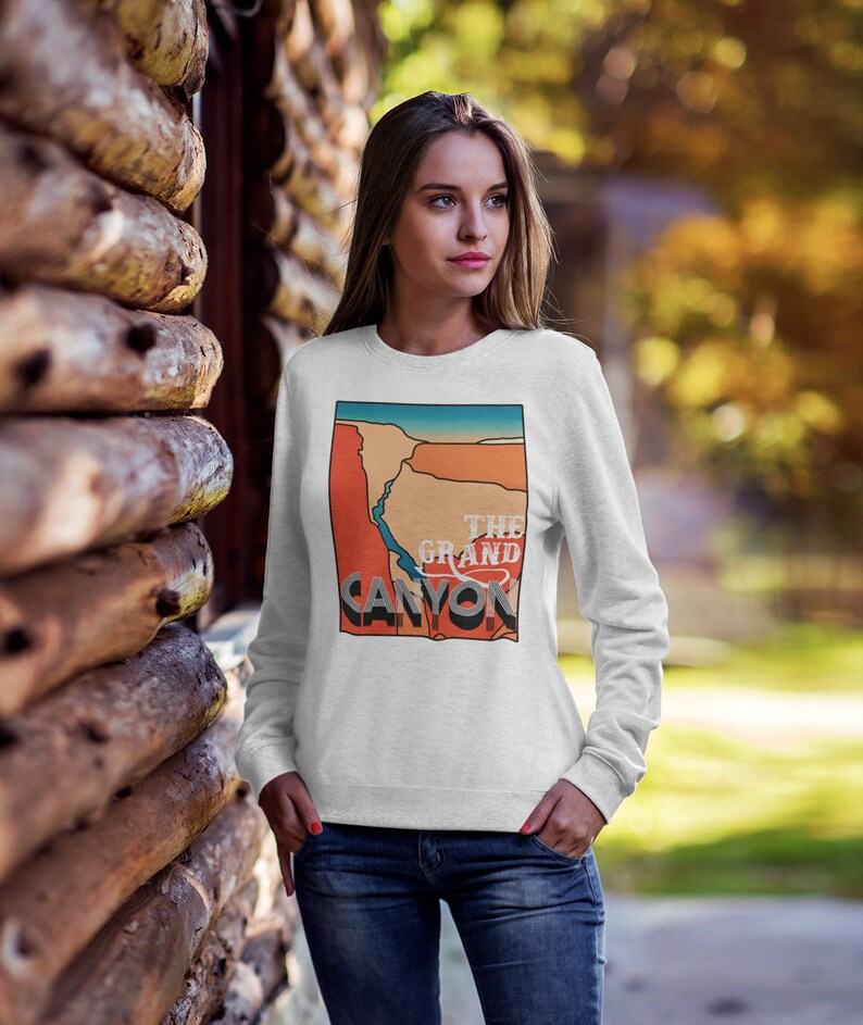 May include: A white sweatshirt featuring a graphic of the Grand Canyon in orange, blue, and beige. The text "THE GRAND CANYON" is printed in a vintage style. The sweatshirt is long-sleeved and worn by a person.