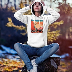 May include: White hoodie with a graphic of the Grand Canyon in red, orange, and blue. The hoodie has a drawstring hood and a front pocket. The text "THE GRAND CANYON" is printed on the graphic. The person is wearing blue jeans and white sneakers.