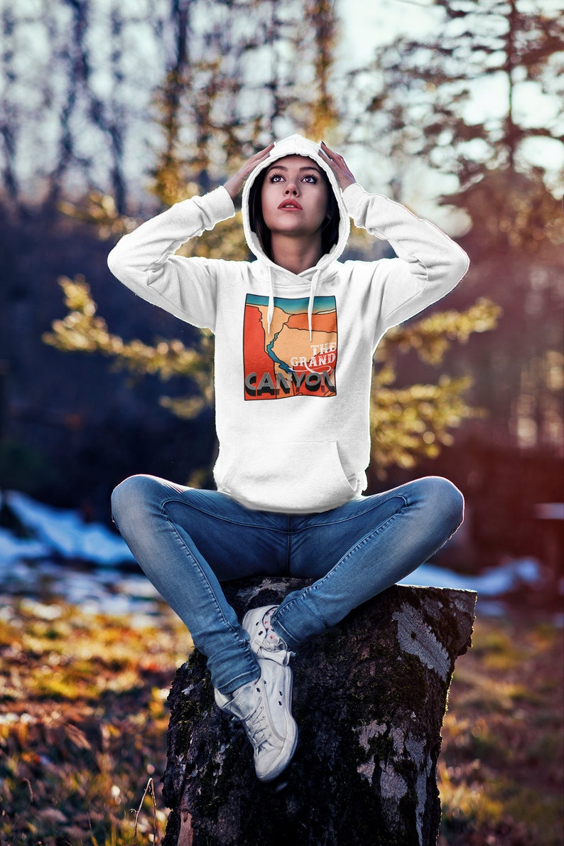 May include: White hoodie with a graphic of the Grand Canyon in red, orange, and blue. The hoodie has a drawstring hood and a front pocket. The text "THE GRAND CANYON" is printed on the graphic. The person is wearing blue jeans and white sneakers.