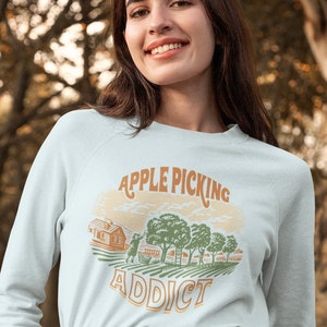 May include: A light blue sweatshirt with a vintage-style graphic of a farmhouse, apple trees, and people picking apples. The text "APPLE PICKING ADDICT" is printed in brown and orange letters.