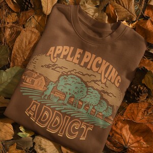 May include: A brown sweatshirt with a vintage-style graphic of a farmhouse, apple trees, and the text "Apple Picking Addict". The sweatshirt is lying on a bed of brown leaves.