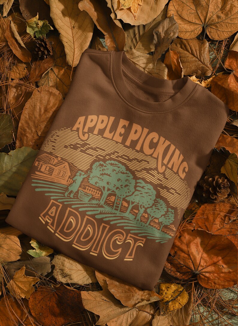 May include: A brown sweatshirt with a vintage-style graphic of a farmhouse, apple trees, and the text "Apple Picking Addict". The sweatshirt is lying on a bed of brown leaves.
