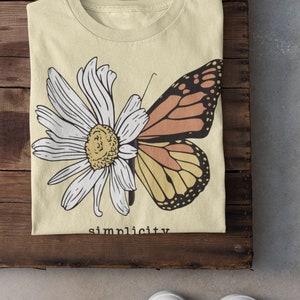 K&ouml;nnte beinhalten: Ein hellgelbes T-Shirt mit einem Motiv aus einer wei&szlig;en Margerite und einem Schmetterling mit orangefarbenen und gelben Fl&uuml;geln. Der Text "simplicity" ist unter dem Motiv aufgedruckt.
