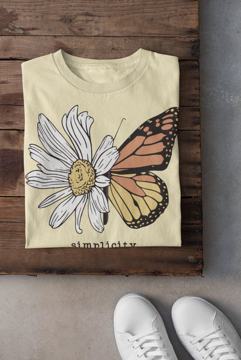 K&ouml;nnte beinhalten: Ein hellgelbes T-Shirt mit einem Motiv aus einer wei&szlig;en Margerite und einem Schmetterling mit orangefarbenen und gelben Fl&uuml;geln. Der Text "simplicity" ist unter dem Motiv aufgedruckt.