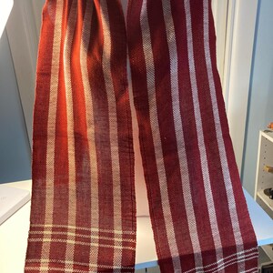 May include: A red and white striped scarf with a woven pattern. The scarf is made of a soft, textured fabric and has a fringed edge.