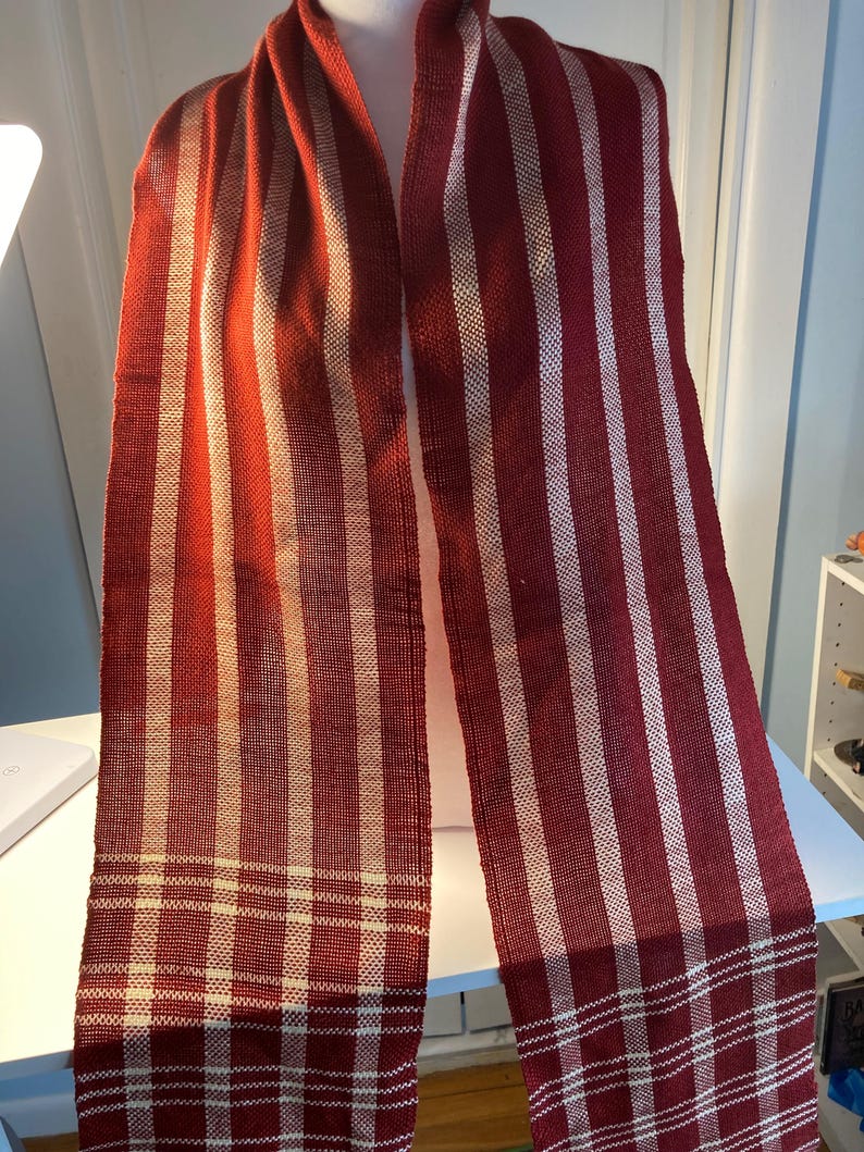 May include: A red and white striped scarf with a woven pattern. The scarf is made of a soft, textured fabric and has a fringed edge.