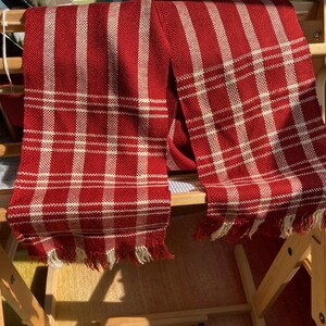 May include: Two red and white plaid scarves with fringe hanging on a wooden loom.