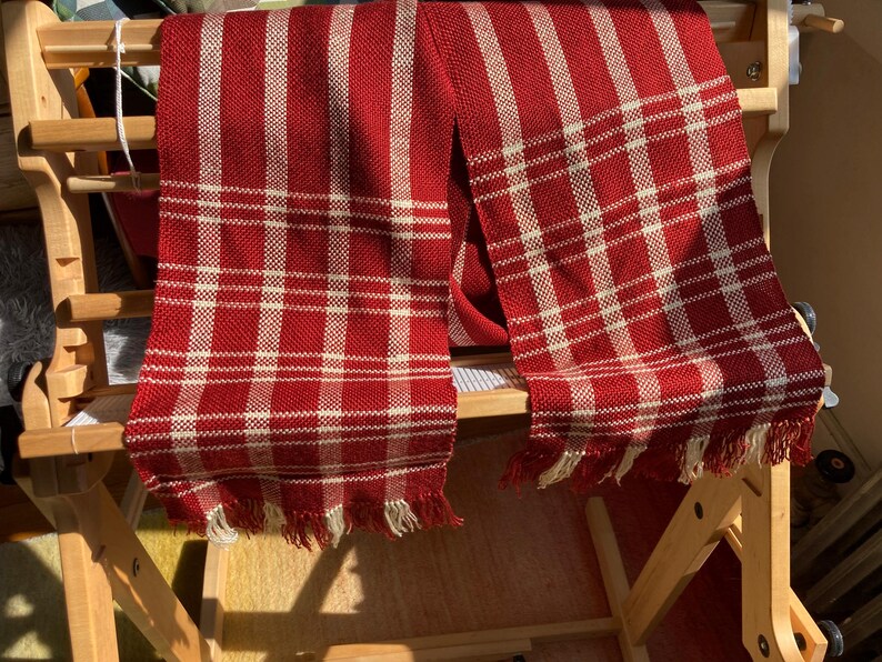 May include: Two red and white plaid scarves with fringe hanging on a wooden loom.
