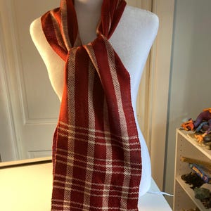 May include: A red and cream striped scarf with a plaid pattern. The scarf is tied around a white mannequin's neck.