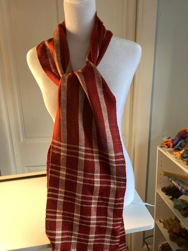 May include: A red and cream striped scarf with a plaid pattern. The scarf is tied around a white mannequin's neck.