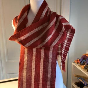 May include: A red and white striped scarf with a fringe on the ends. The scarf is woven with a simple pattern and is made of a soft, natural fiber.
