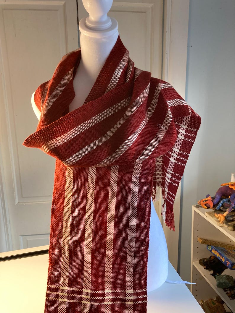 May include: A red and white striped scarf with a fringe on the ends. The scarf is woven with a simple pattern and is made of a soft, natural fiber.