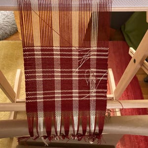 May include: A red and white plaid fabric woven on a wooden loom. The fabric is partially finished and has fringe on the bottom.