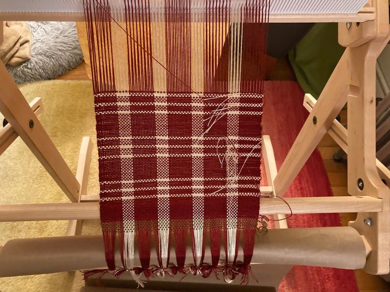 May include: A red and white plaid fabric woven on a wooden loom. The fabric is partially finished and has fringe on the bottom.