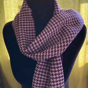 May include: A purple and white houndstooth pattern scarf draped over a black mannequin. The scarf is made of a soft material and has a classic design. The scarf is a winter accessory.