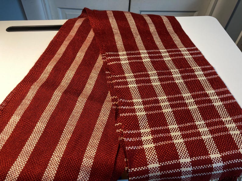 May include: A red and white striped woven fabric with a red and white plaid pattern. The fabric is folded in half and is lying on a white surface.