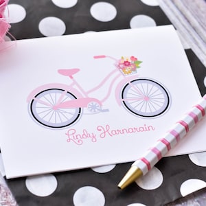 May include: A pink bicycle with a basket of flowers on the handlebars. The bicycle is on a white card with the name "Lindy Harnarain" written in pink cursive.