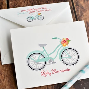 May include: A light blue bicycle with a basket full of flowers. The bicycle has a white handlebar and white tires. The name "Lindy Harnarain" is written in red cursive below the bicycle.