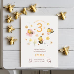May include: A birthday invitation with the text "So Sweet to Bee" and "3" in orange. The invitation features illustrations of bees and flowers. Decorative bee figurines surround the invitation.