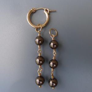 Puede incluir: Un par de pendientes de aro de oro con gotas de perlas negras. Cada pendiente tiene tres perlas negras que cuelgan del aro.