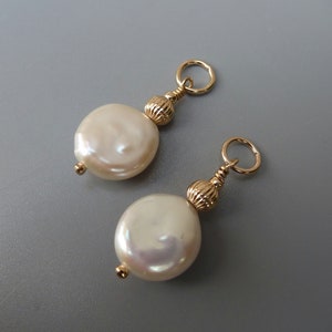 Puede incluir: Dos colgantes dorados para pendientes con perlas grandes, redondas y planas, de color blanquecino. Cada colgante tiene una pequeña cuenta dorada en la parte inferior de la perla, una cuenta dorada texturizada y un lazo dorado en la parte superior. El fondo es gris liso.