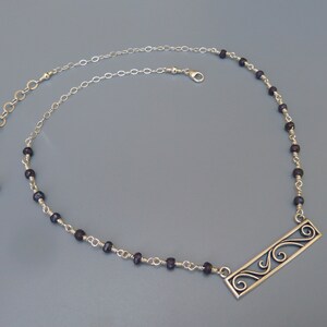 Op de afbeelding: Een zilveren ketting met een zilveren hanger met een decoratief ontwerp. De ketting is gemaakt met kleine, donkerpaarse kralen en een zilveren ketting.