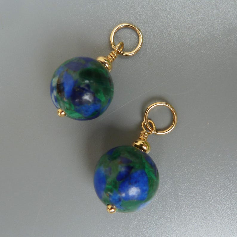 May include: A pair of earrings featuring round, marbled beads in shades of blue and green. Each bead is accented with gold-colored metal findings, including a small loop and a decorative cap. The earrings are displayed against a neutral gray background.