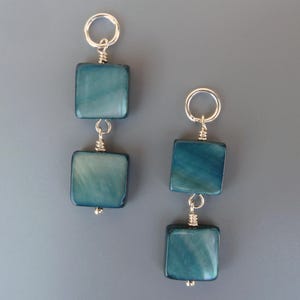 Puede incluir: Un par de pendientes cuadrados de color turquesa. Cada pendiente presenta dos cuentas cuadradas, iridiscentes y turquesas, conectadas por alambre de plata. Los pendientes tienen un aro plateado en la parte superior para colgar.
