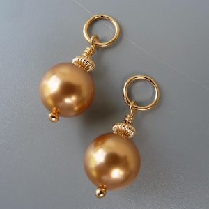 Puede incluir: Dos abalorios de perlas doradas con un anillo dorado en la parte superior. Las perlas son redondas y tienen un acabado liso y brillante.