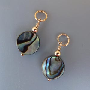 Puede incluir: Dos abalorios dorados con cuentas redondas de concha de abulón. Las cuentas tienen patrones iridiscentes azules, verdes y negros.