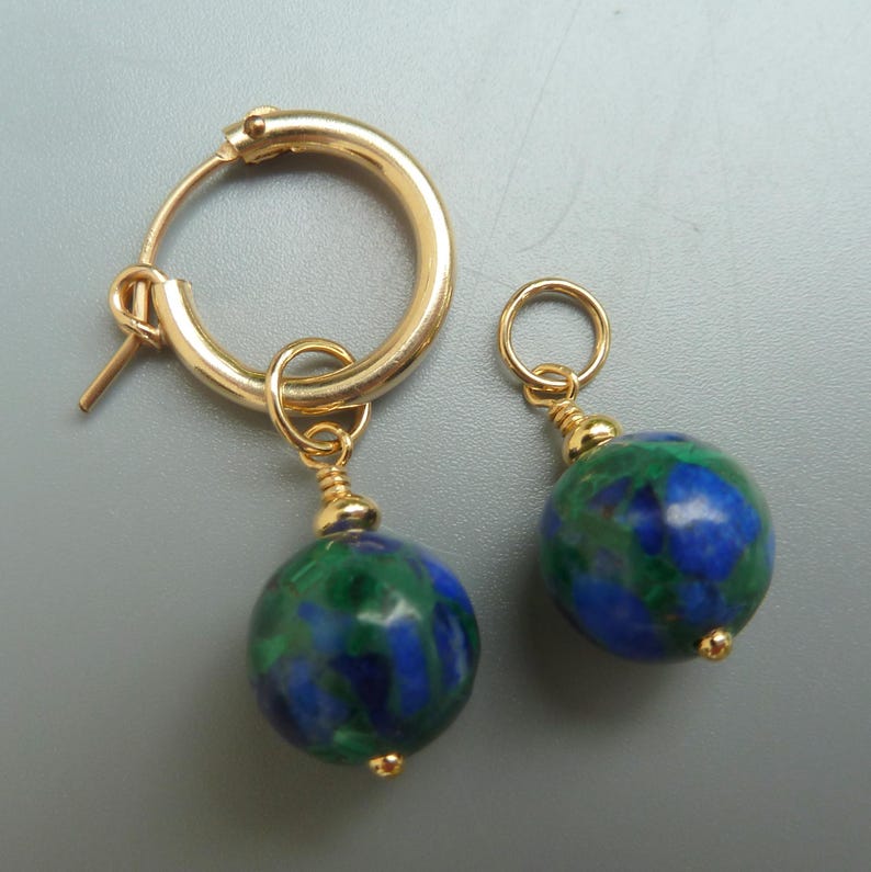 May include: A pair of gold-colored hoop earrings with a round, blue and green stone pendant. The earrings have a simple design with a small gold bead at the bottom of the pendant. The earrings are displayed on a gray background.