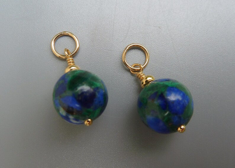 May include: Two round earrings with gold-colored hoops and accents. Each earring features a blue and green marbled stone. The earrings are displayed against a gray background.