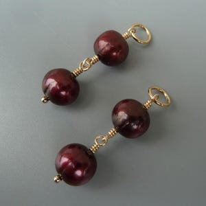 Puede incluir: Un par de pendientes con dos perlas de color rojo oscuro en cada pendiente, conectadas por un alambre de oro. Los pendientes tienen un aro de salto de oro en la parte superior.