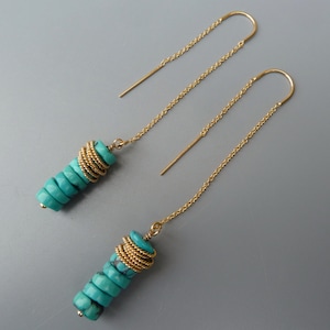 May include: A pair of turquoise and gold dangle earrings. The earrings feature a stack of turquoise beads with gold wire wrapped around them. The earrings are suspended from a thin gold chain.
