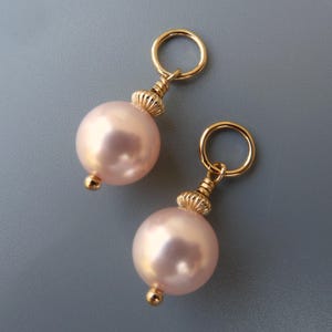 Puede incluir: Dos abalorios dorados con una perla rosa unida a cada uno. Los abalorios tienen un pequeño anillo dorado en la parte superior.