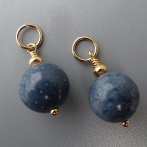 Puede incluir: Dos abalorios de cuentas de piedras preciosas azules y doradas con anillas de salto doradas. Las cuentas son redondas y tienen una textura moteada.